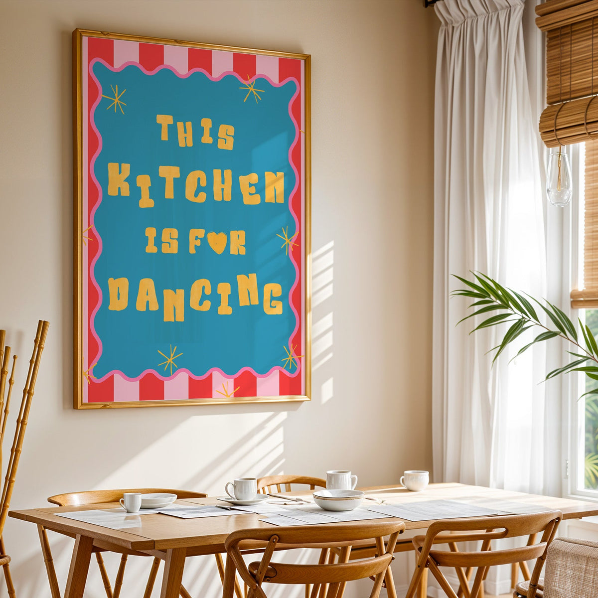 This Kitchen Is For Dancing print shown in a gold frame above a breakfast table by a curtained window