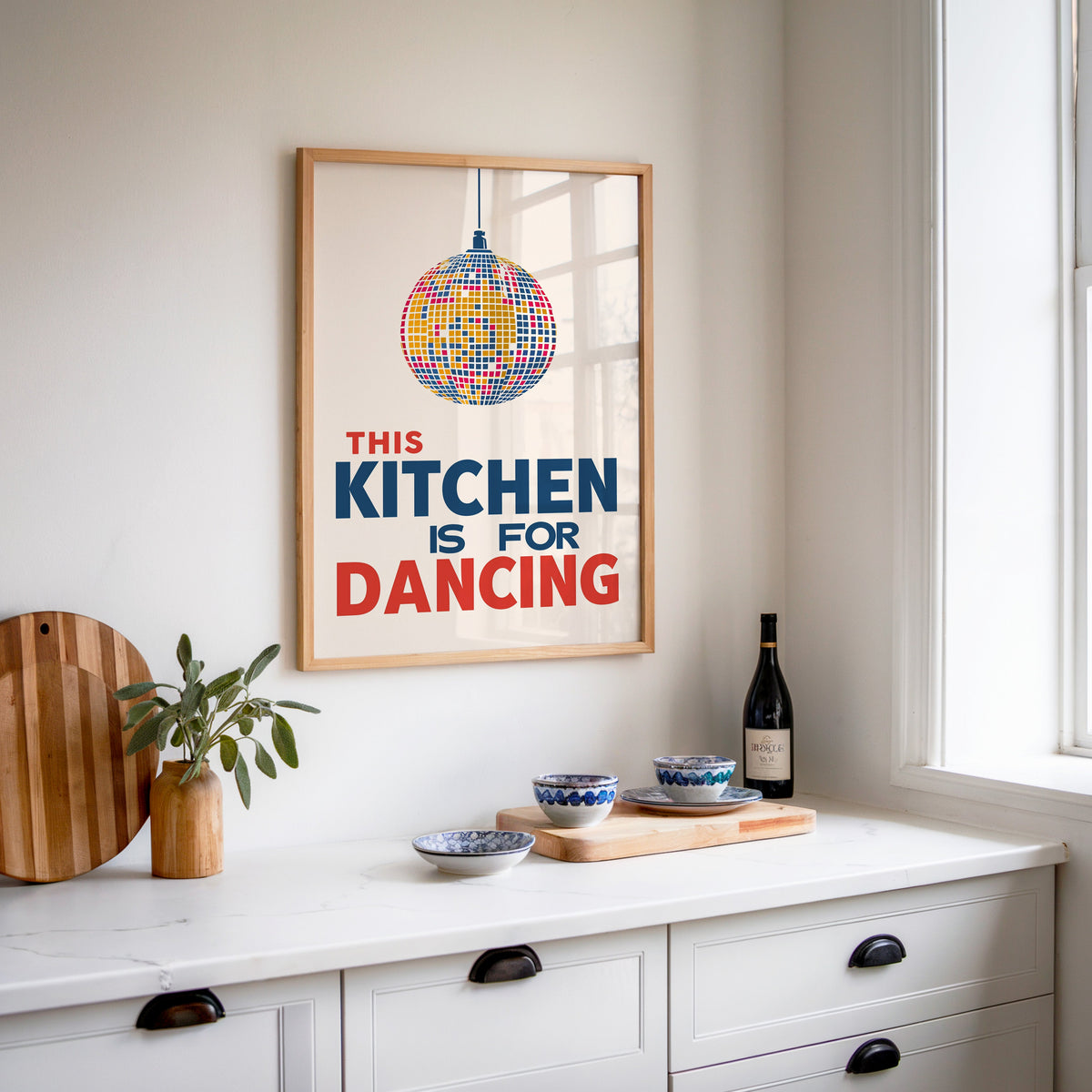 Kitchen Is For Dancing blue disco print in an oak frame above a white sideboard with a bottle of wine