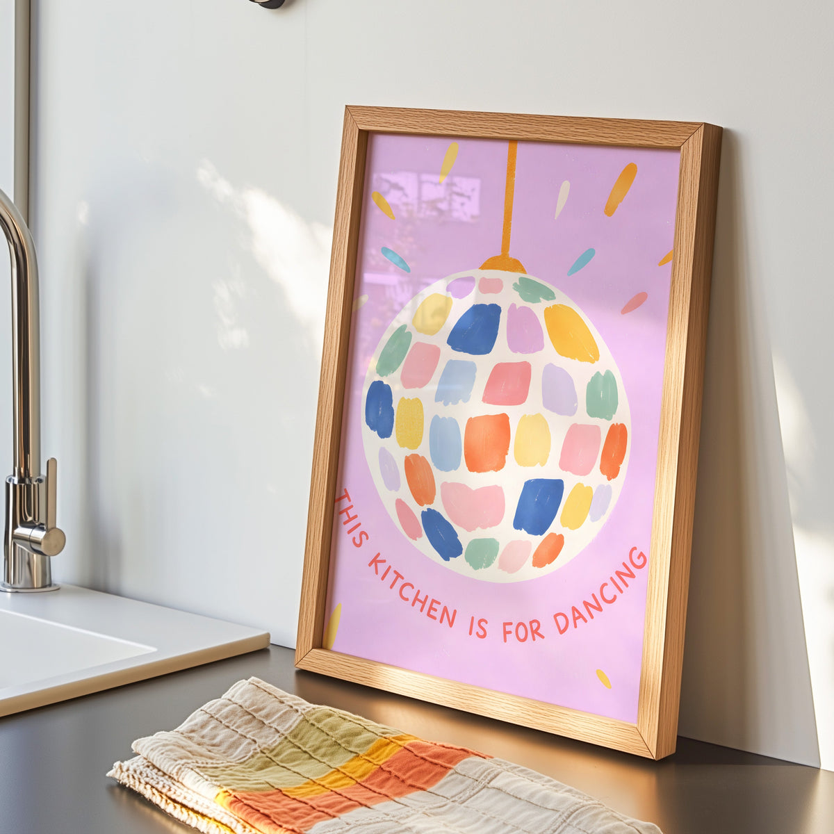 Pastel disco ball print in an oak frame by the kitchen sink with a striped tea towel