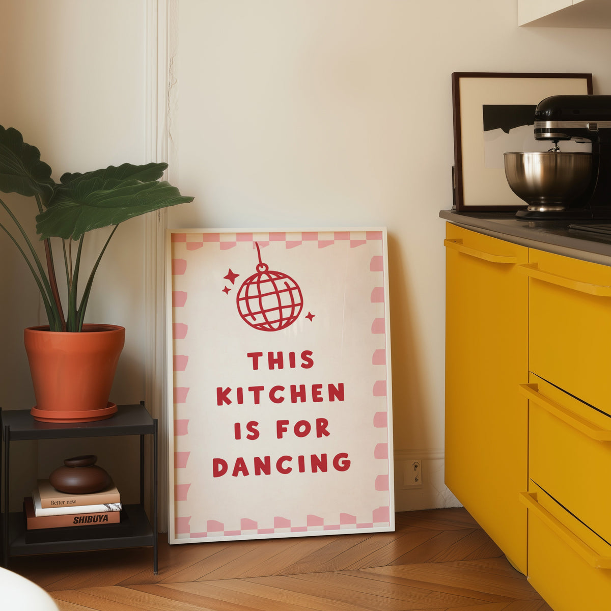 Kitchen Is For Dancing pink check print in a white frame leaning on parquet beside a bright yellow kitchen