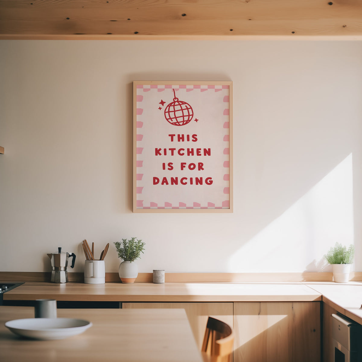 Kitchen Is For Dancing pink check print in an oak frame above a wood counter with a moka pot and herbs