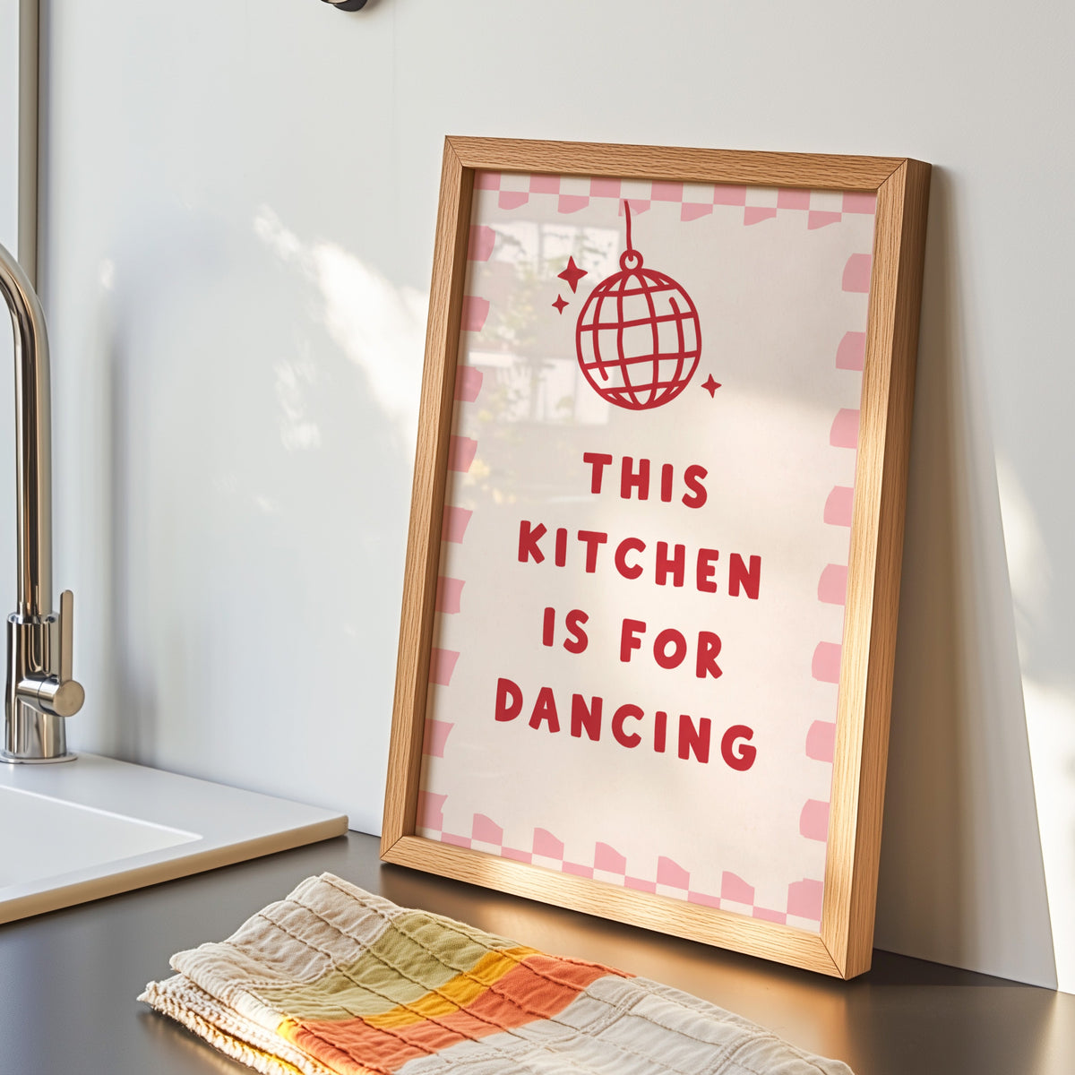 Kitchen Is For Dancing pink check print in an oak frame by the kitchen sink beside a striped tea towel