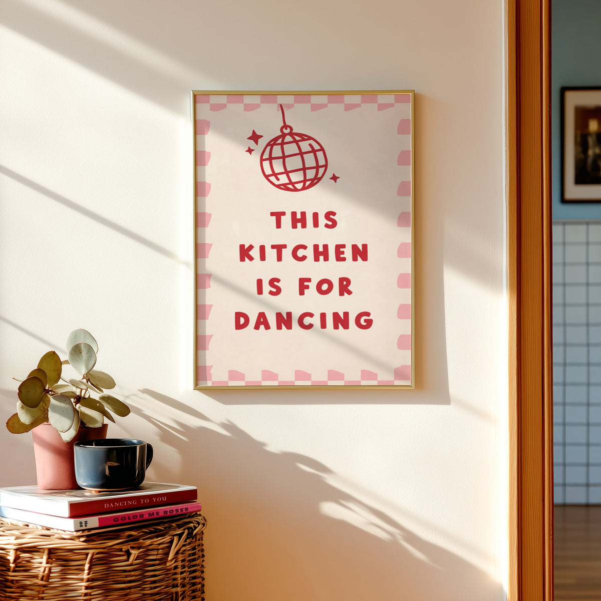 Kitchen Is For Dancing pink check print shown in a gold frame above a basket of books and a pilea plant