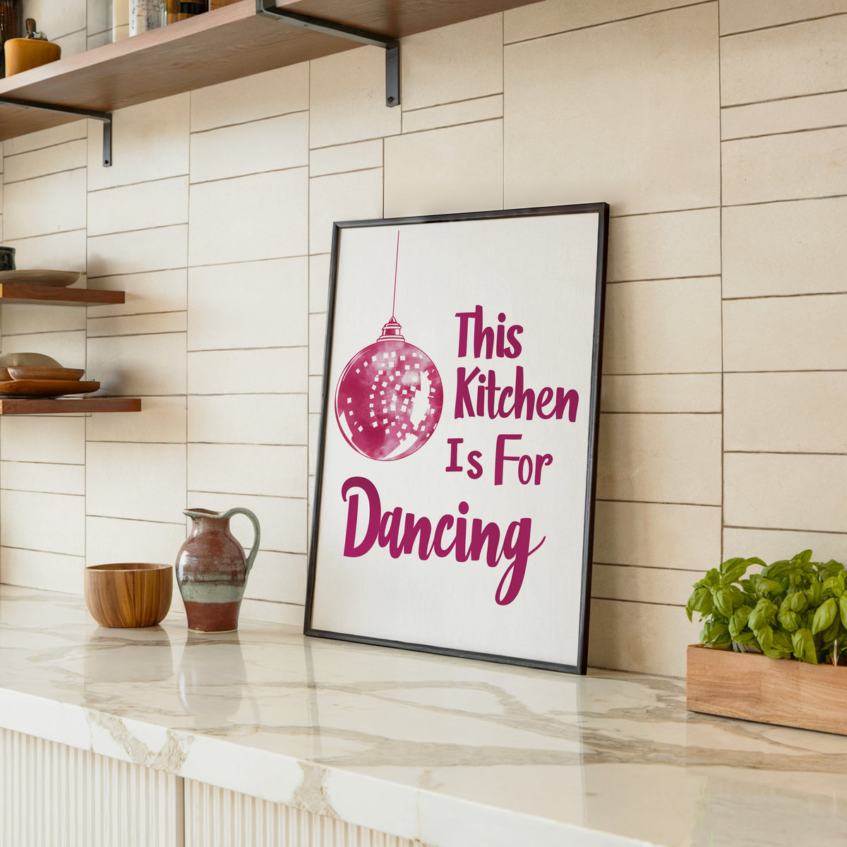 Kitchen Is For Dancing pink script print in a black frame leaning on a marble counter in a tiled kitchen