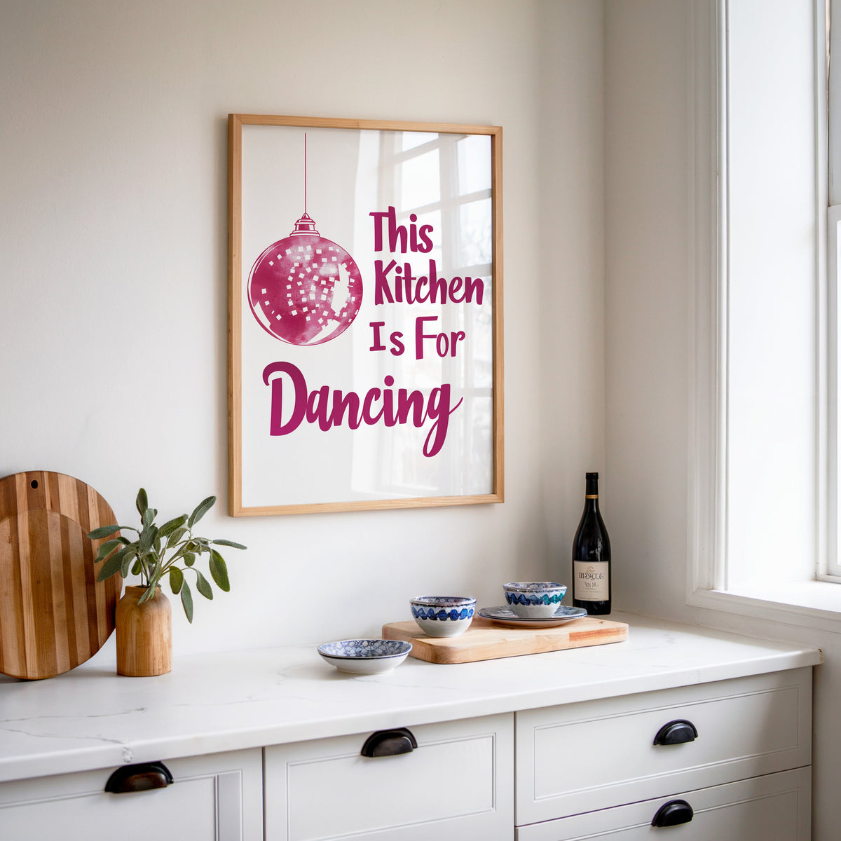 Kitchen Is For Dancing pink script print in an oak frame above a white sideboard with a bottle of wine