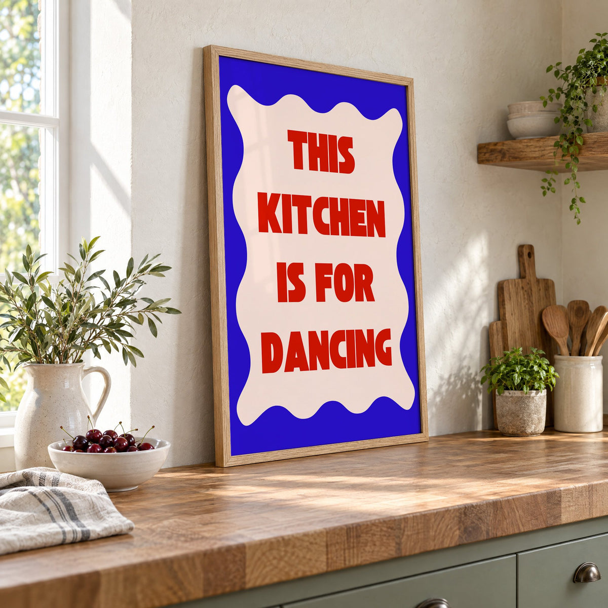 Kitchen Is for Dancing wavy blue print in an oak frame leaning on a butcher block island beside a bowl of cherries
