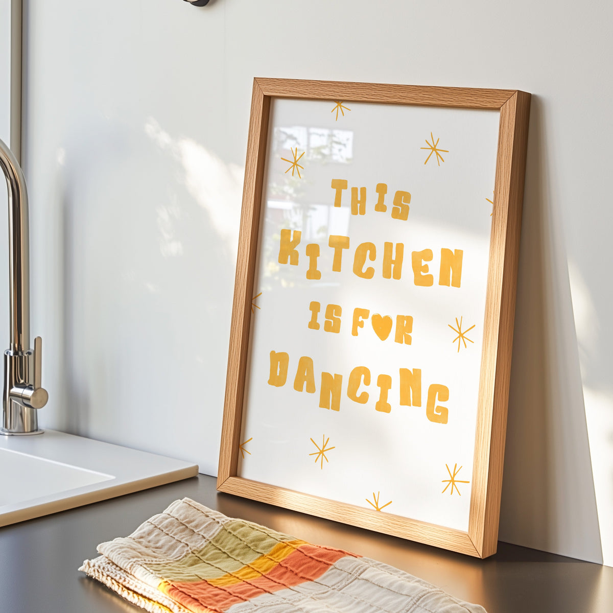 Kitchen Is For Dancing yellow star print in an oak frame by the kitchen sink beside a striped tea towel