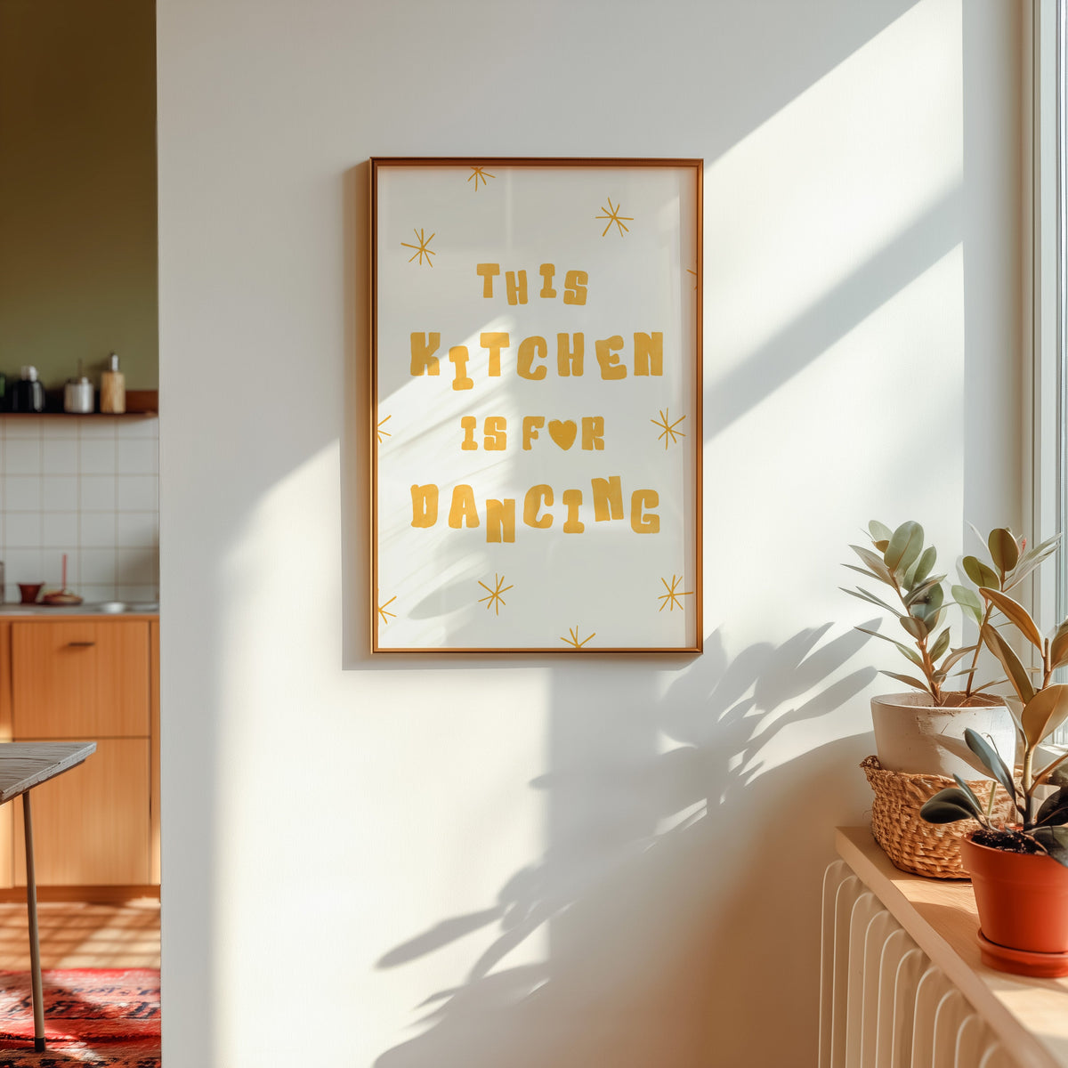 Kitchen Is For Dancing yellow star print shown in a gold frame in sunlight with plants and a kitchen beyond
