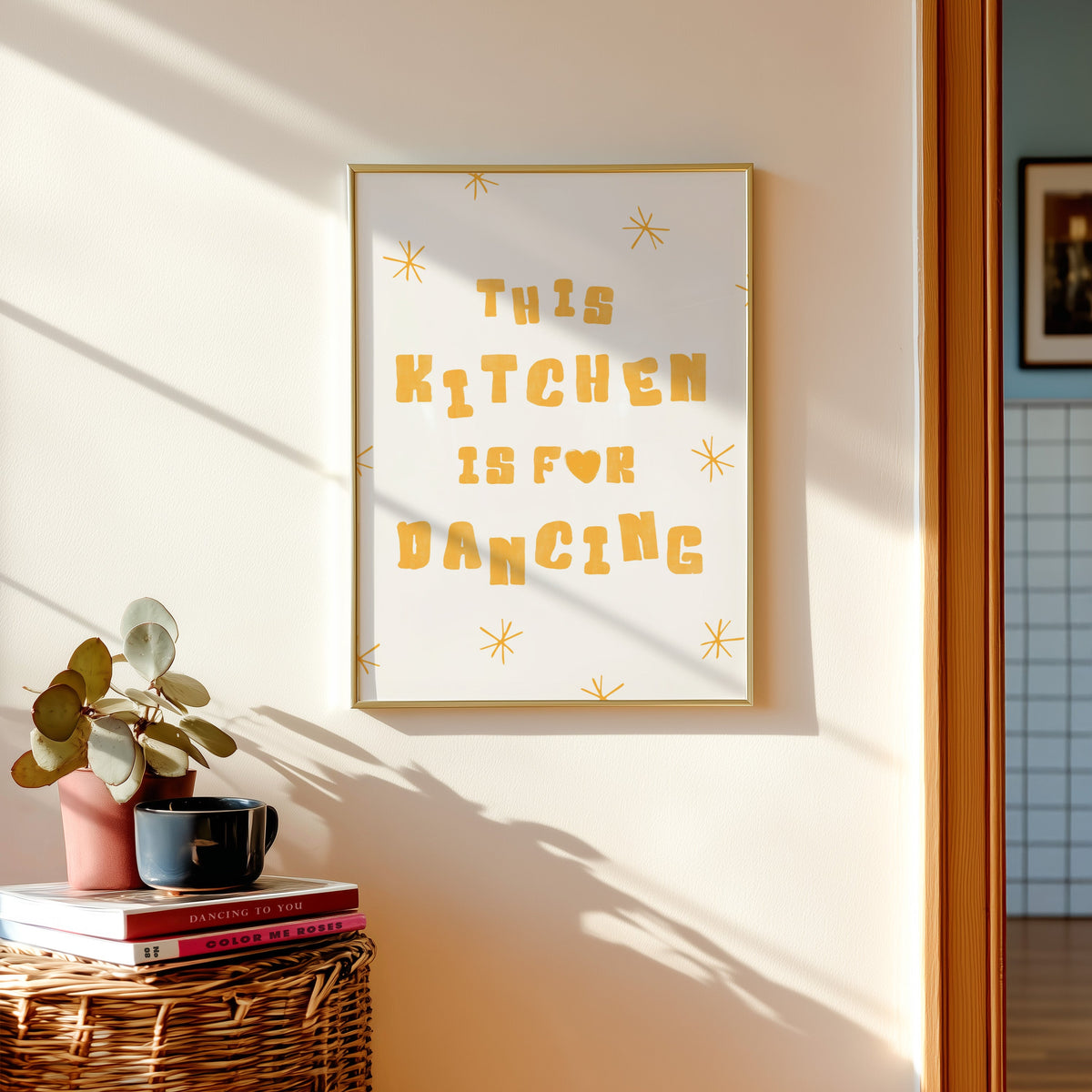Kitchen Is For Dancing yellow star print shown in a gold frame above a basket of books and a pilea plant