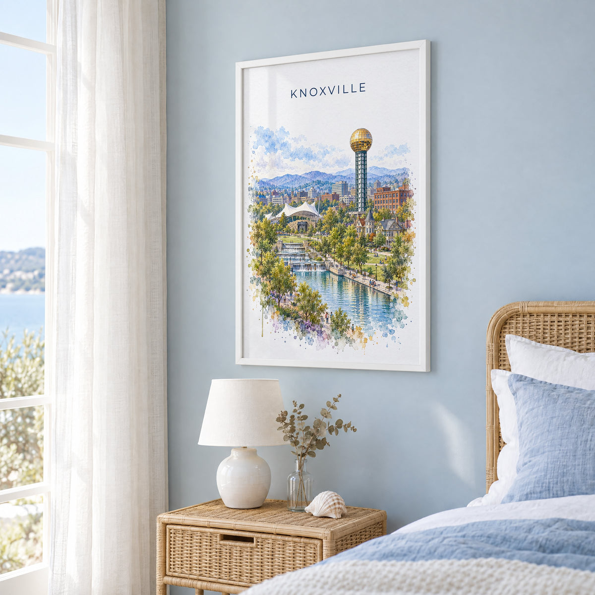 Knoxville watercolor print in a white frame on a pale blue wall beside a rattan bed in a coastal bedroom