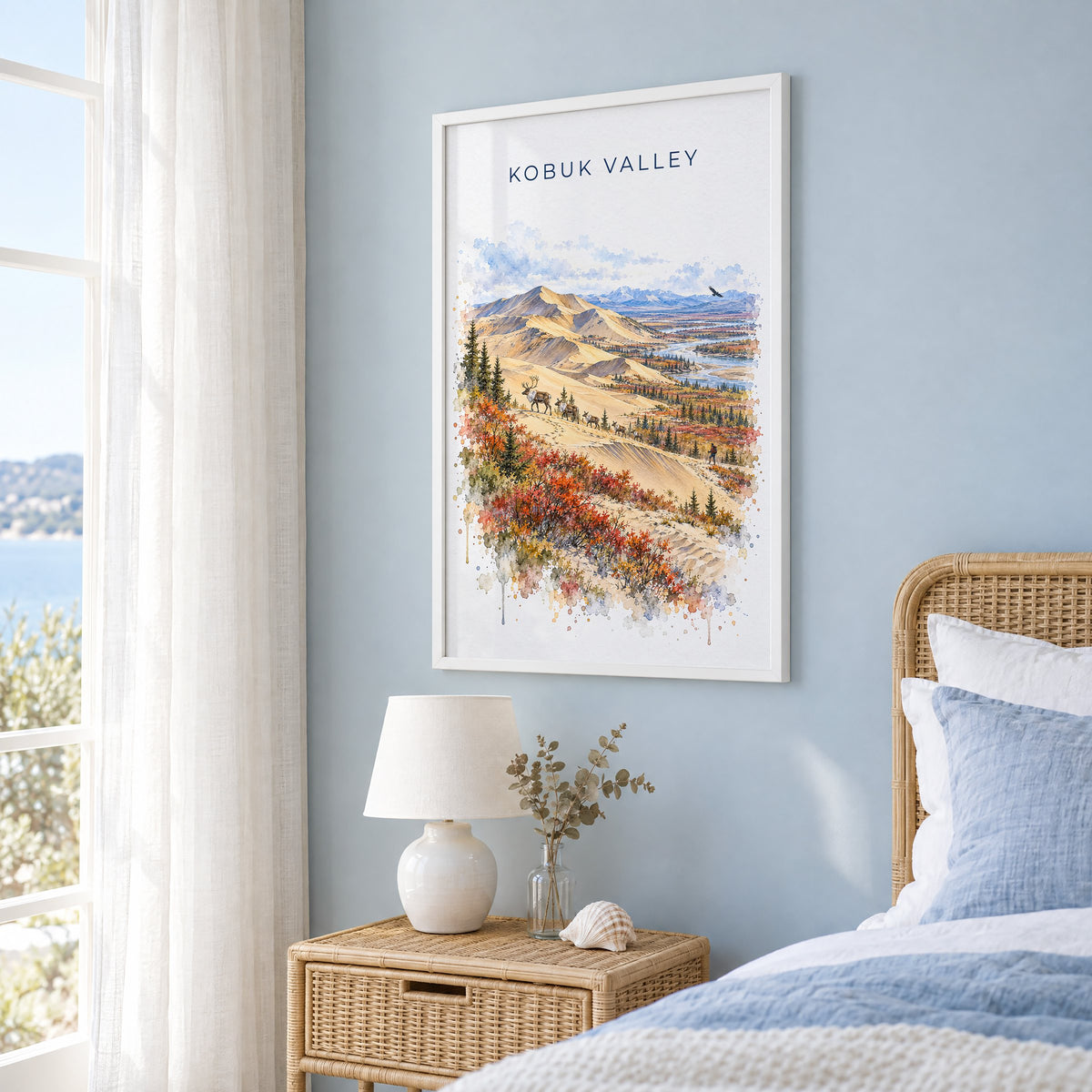 Kobuk Valley National Park watercolor print in a white frame on a pale blue wall beside a rattan bed in a coastal bedroom