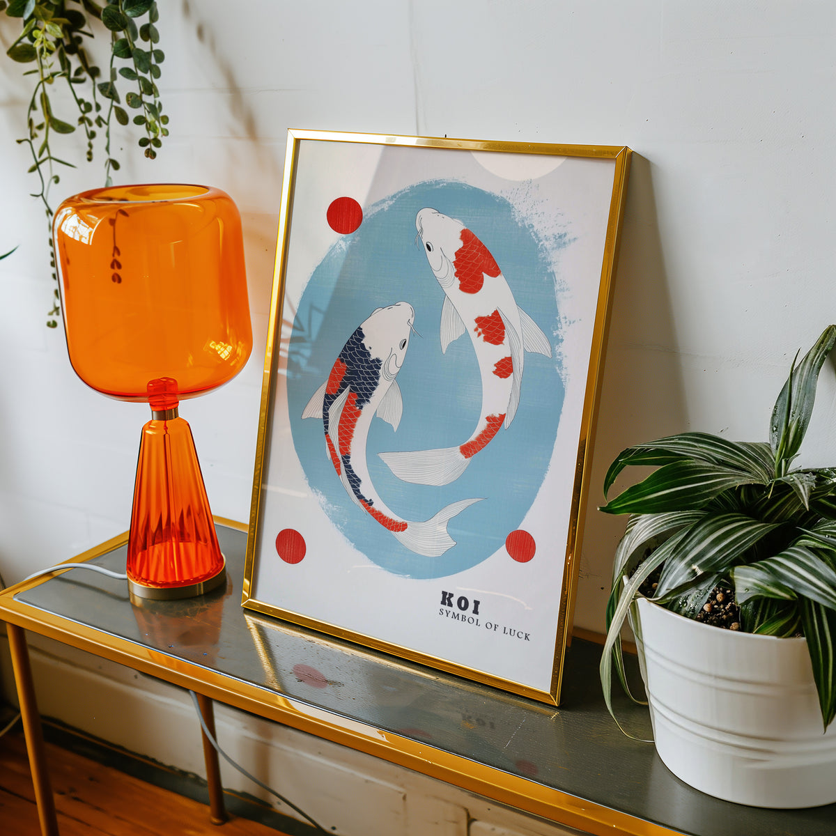 Two koi circling in blue oval print shown in a gold frame on a console beside an orange glass lamp