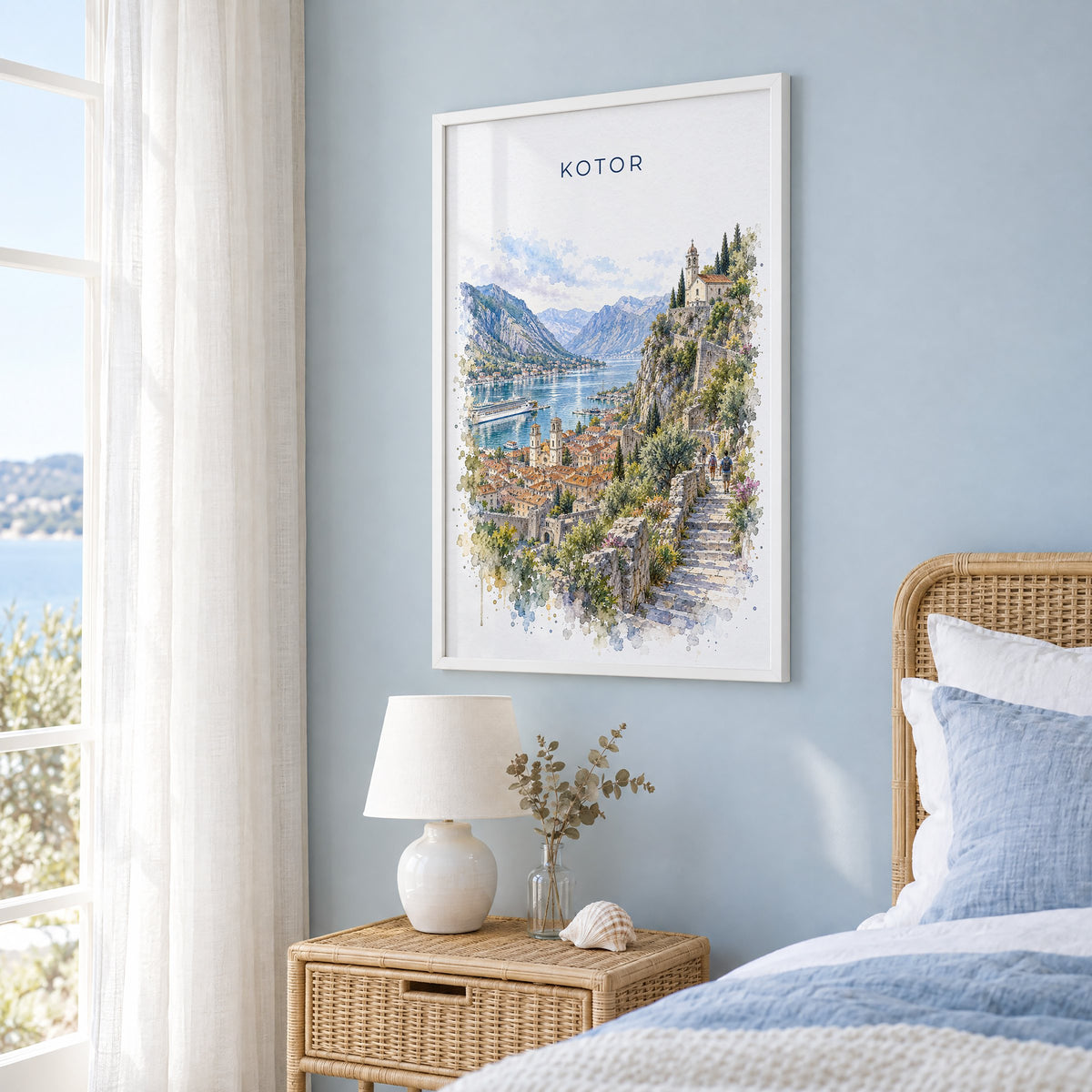 Kotor watercolor print in a white frame on a pale blue wall beside a rattan bed in a coastal bedroom