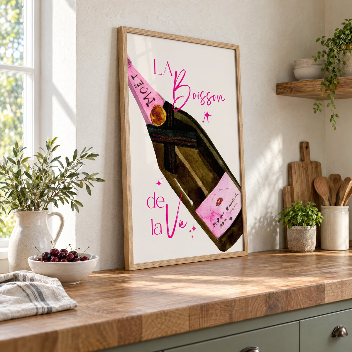 La Boisson champagne bottle watercolor print in an oak frame leaning on a butcher block island beside a bowl of cherries