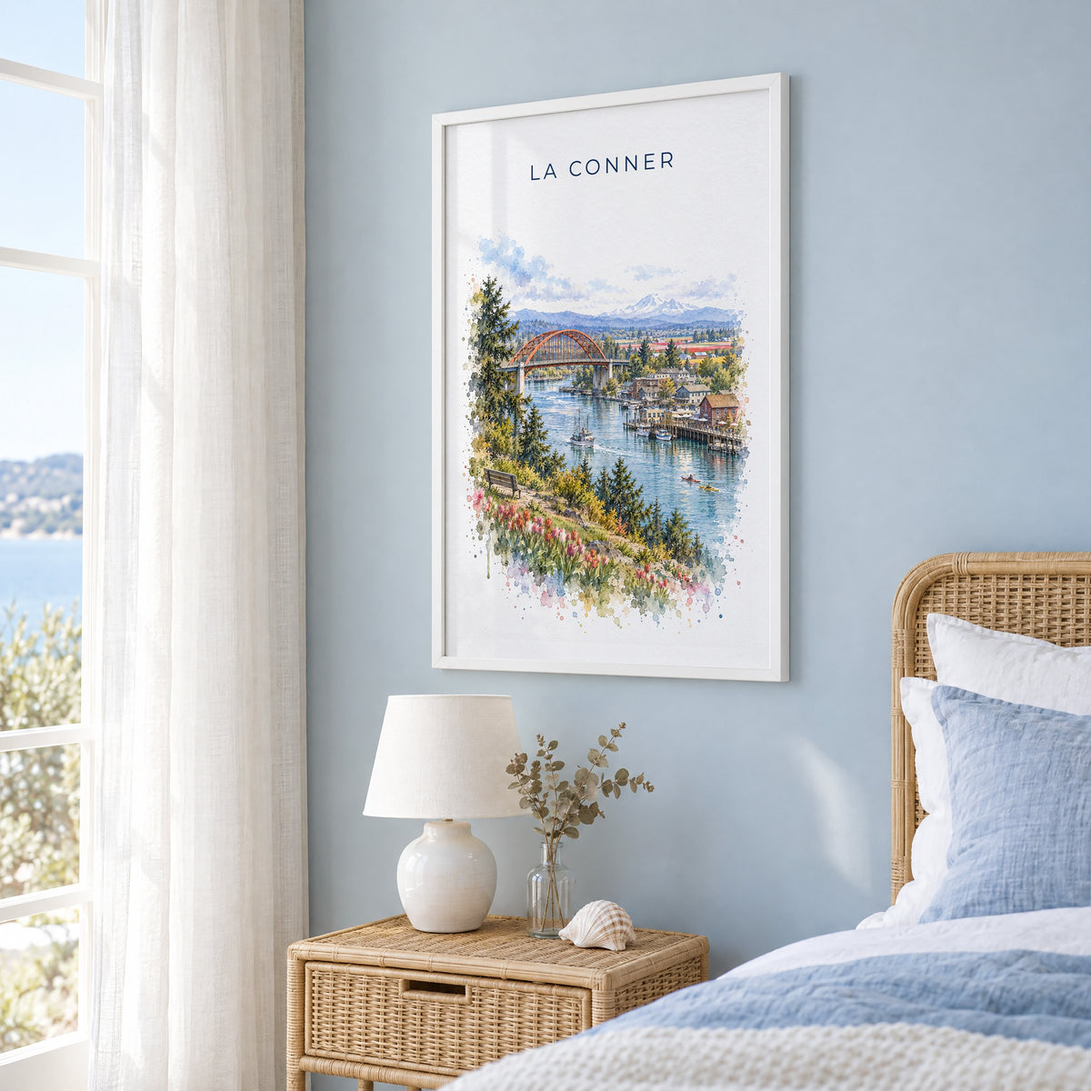 La Conner, Washington watercolor print in a white frame on a pale blue wall beside a rattan bed in a coastal bedroom