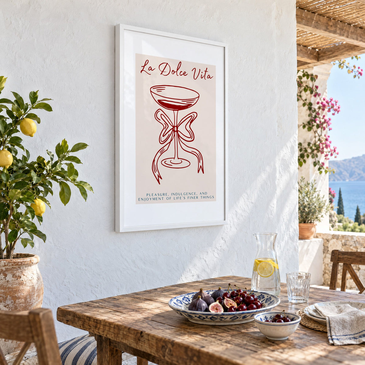 La Dolce Vita coupe bow line print in a white frame on a whitewashed terrace wall above a lunch table with figs and cherries, seen at an angle