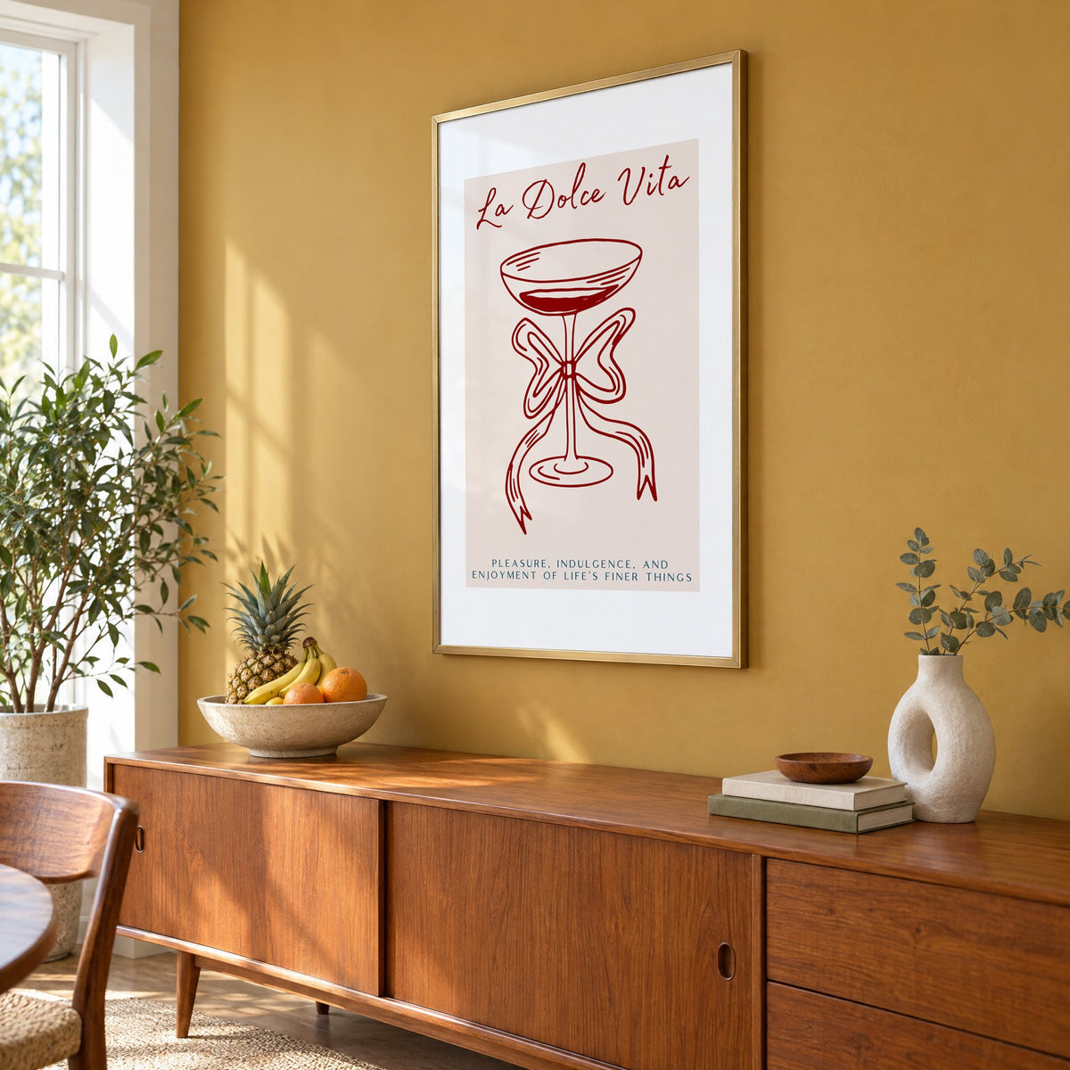 La Dolce Vita coupe bow line print in a brass frame on a mustard wall above a teak sideboard with a fruit bowl, seen at an angle
