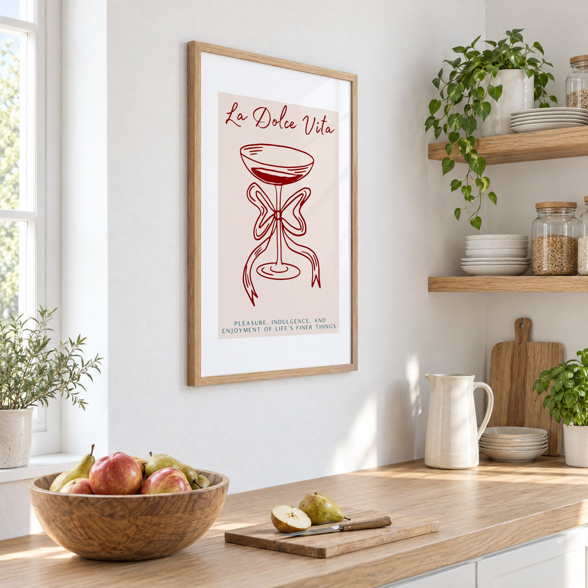 La Dolce Vita coupe bow line print in an oak frame on a white kitchen wall above a counter with apples and pears, seen at an angle
