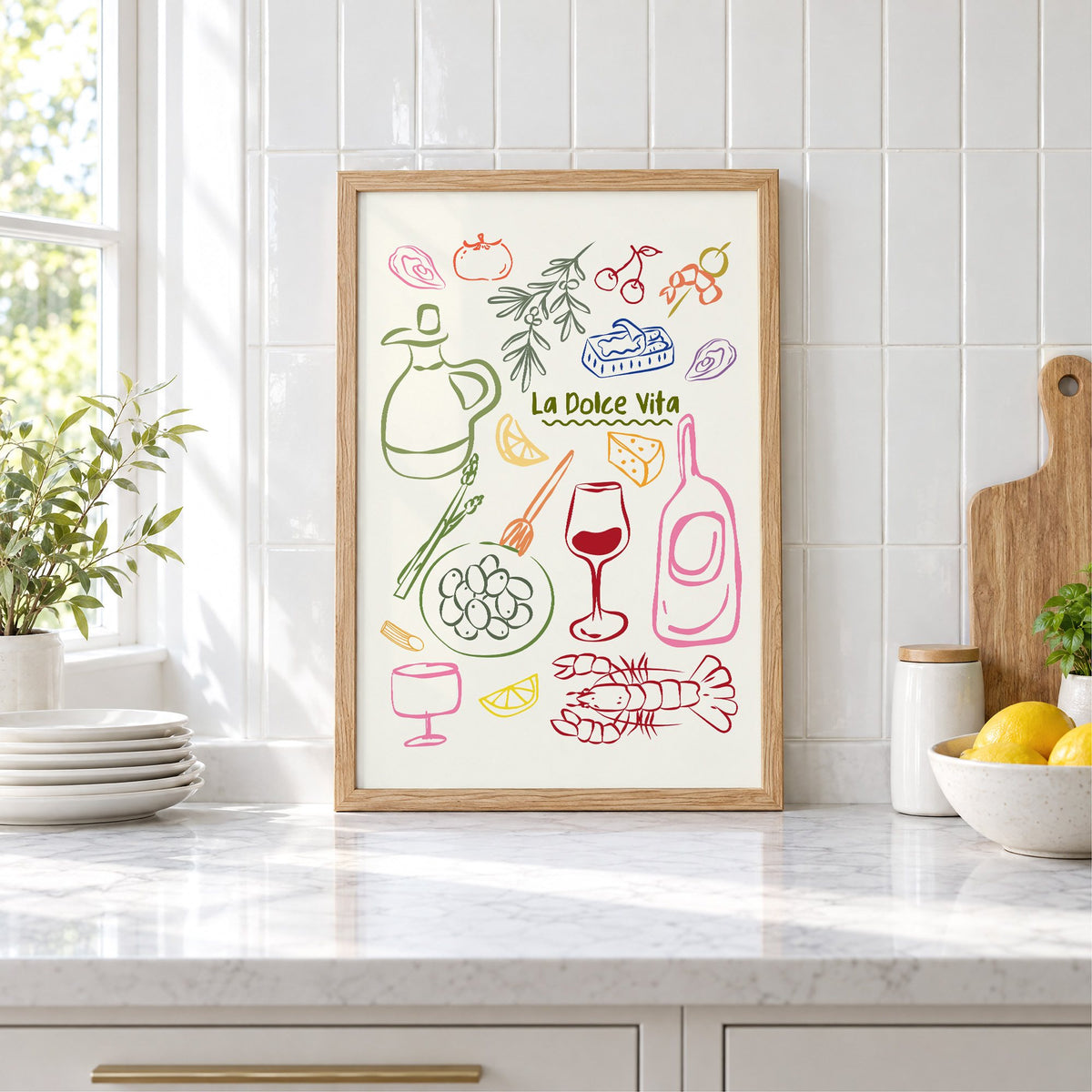 La Dolce Vita feast doodle print in an oak frame leaning on a marble counter by white tiles and lemons