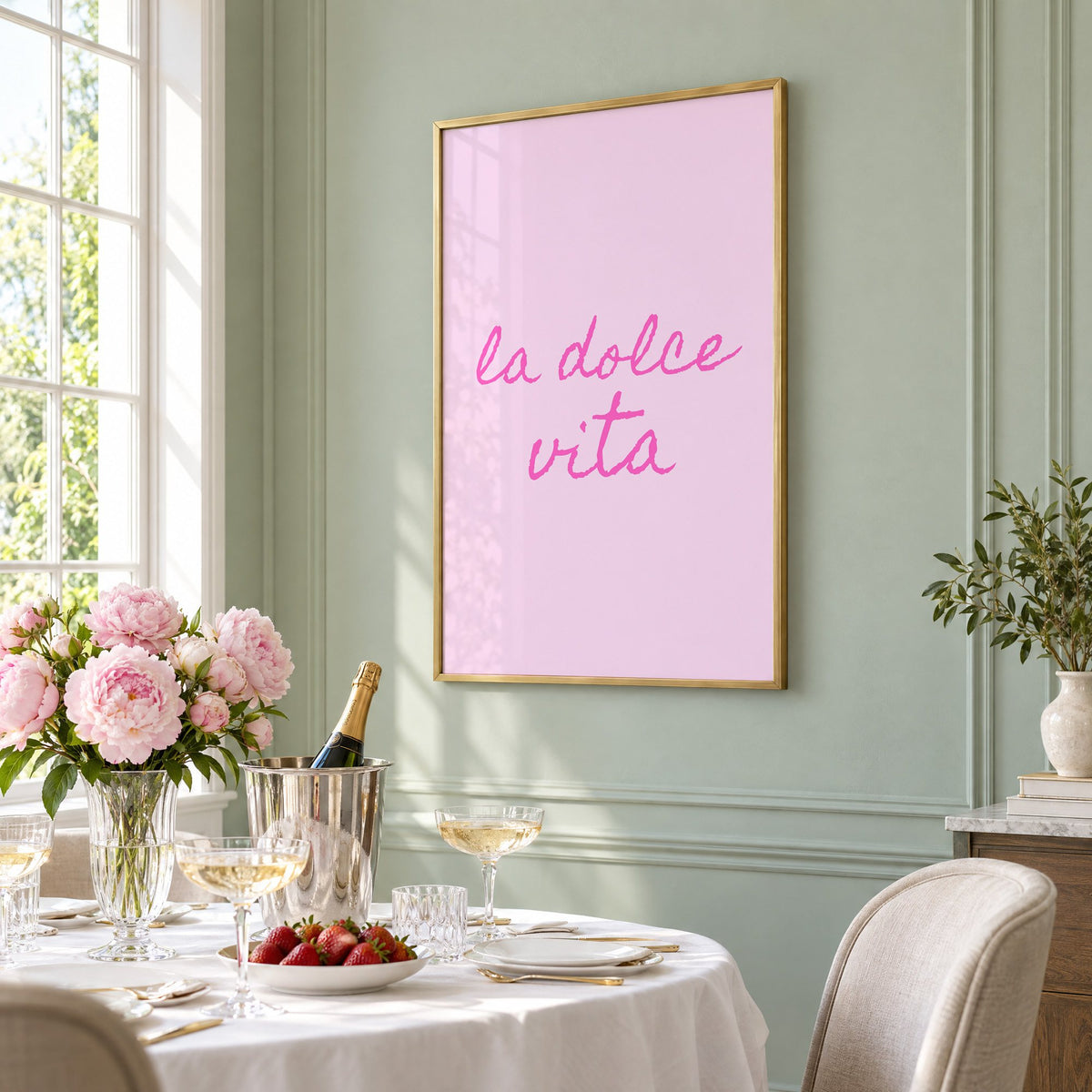 la dolce vita pink handwritten script print in a brass frame on a sage wall above a brunch table with champagne and peonies, seen at an angle