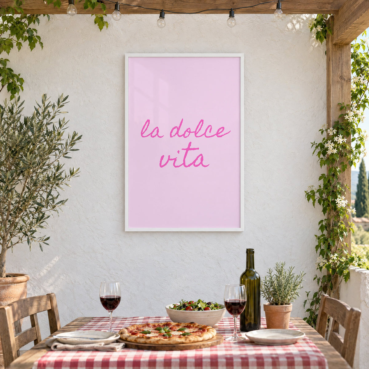 la dolce vita pink handwritten script print in a white frame on a stucco wall above a checked-cloth table with pizza and red wine