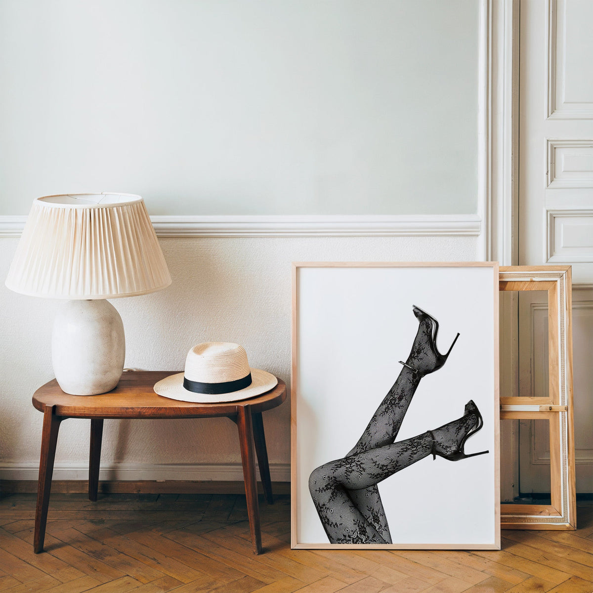 Legs in floral lace tights and stilettos print in a pine frame on a wood floor beside a lamp and straw hat