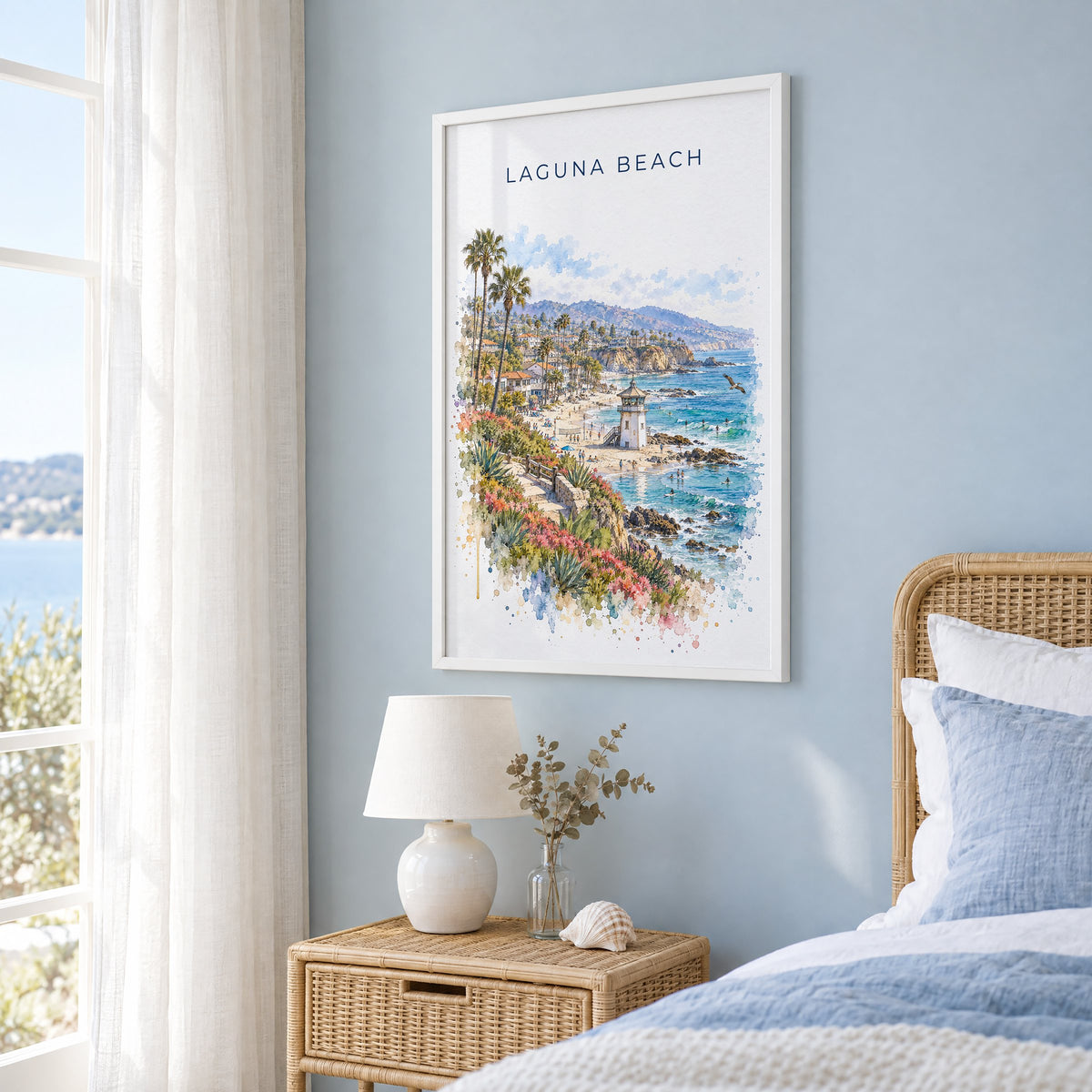 Laguna Beach watercolor print in a white frame on a pale blue wall beside a rattan bed in a coastal bedroom