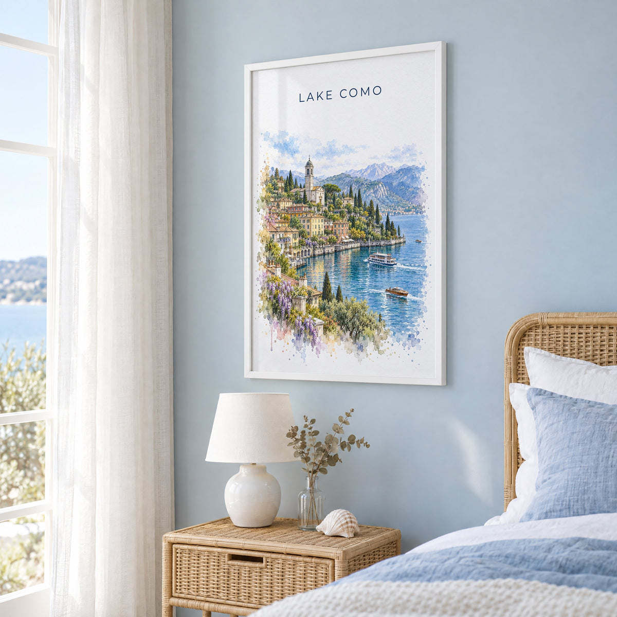 Lake Como watercolor print in a white frame on a pale blue wall beside a rattan bed in a coastal bedroom