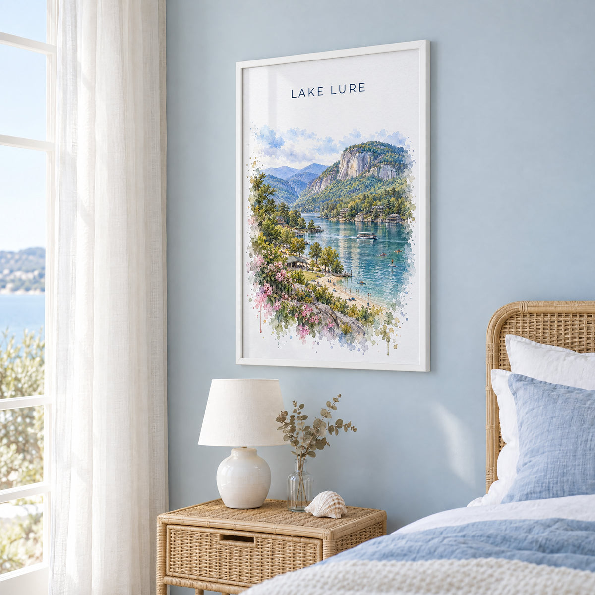 Lake Lure watercolor print in a white frame on a pale blue wall beside a rattan bed in a coastal bedroom