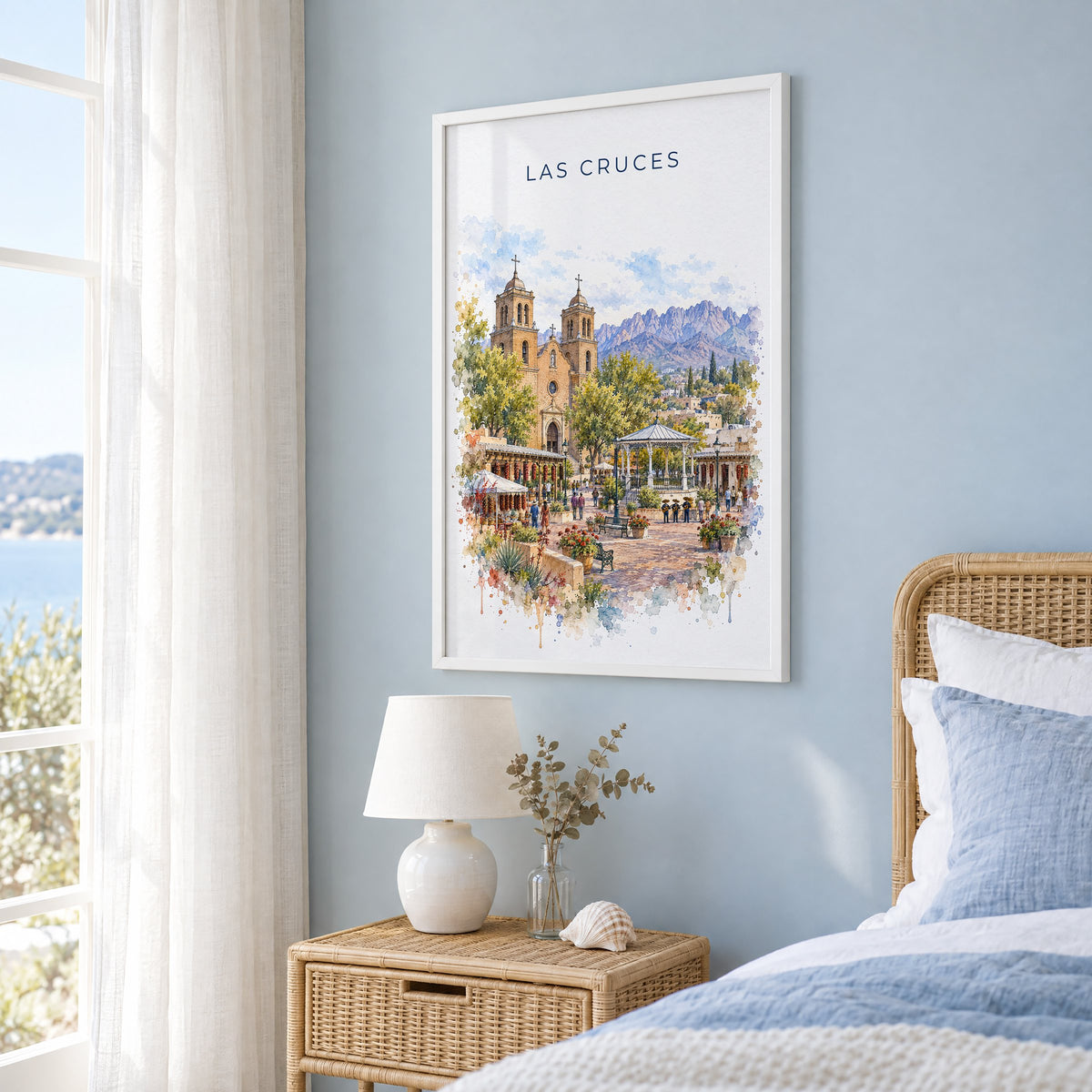 Las Cruces watercolor print in a white frame on a pale blue wall beside a rattan bed in a coastal bedroom