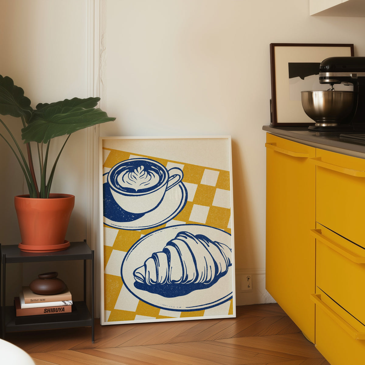 Latte and croissant print in a white frame leaning on parquet beside a bright yellow kitchen