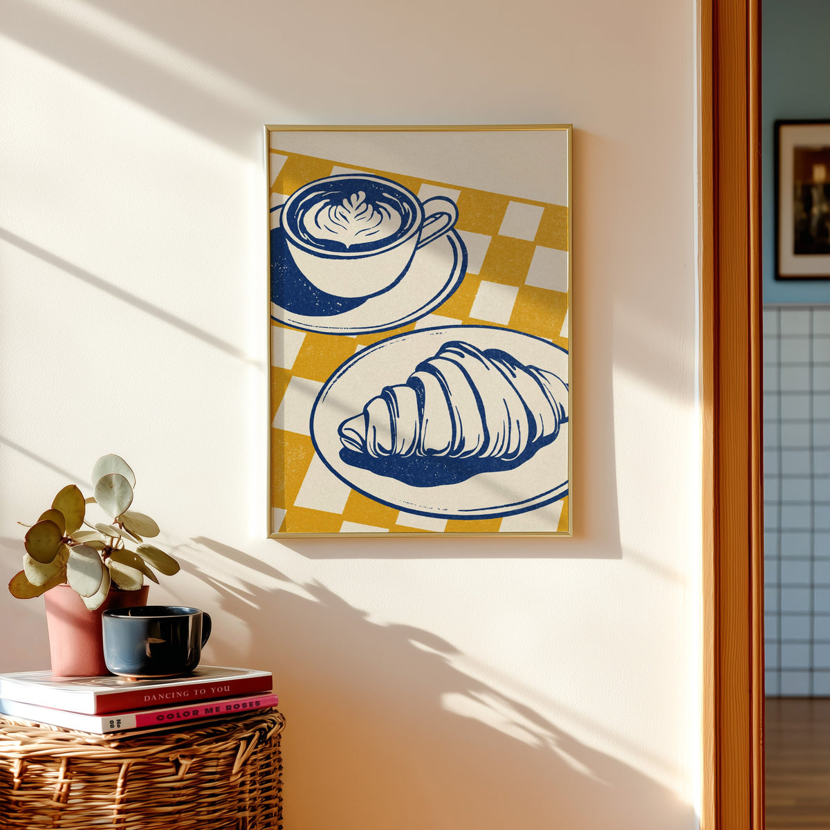 Latte and croissant print shown in a gold frame above a basket of books and a pilea plant