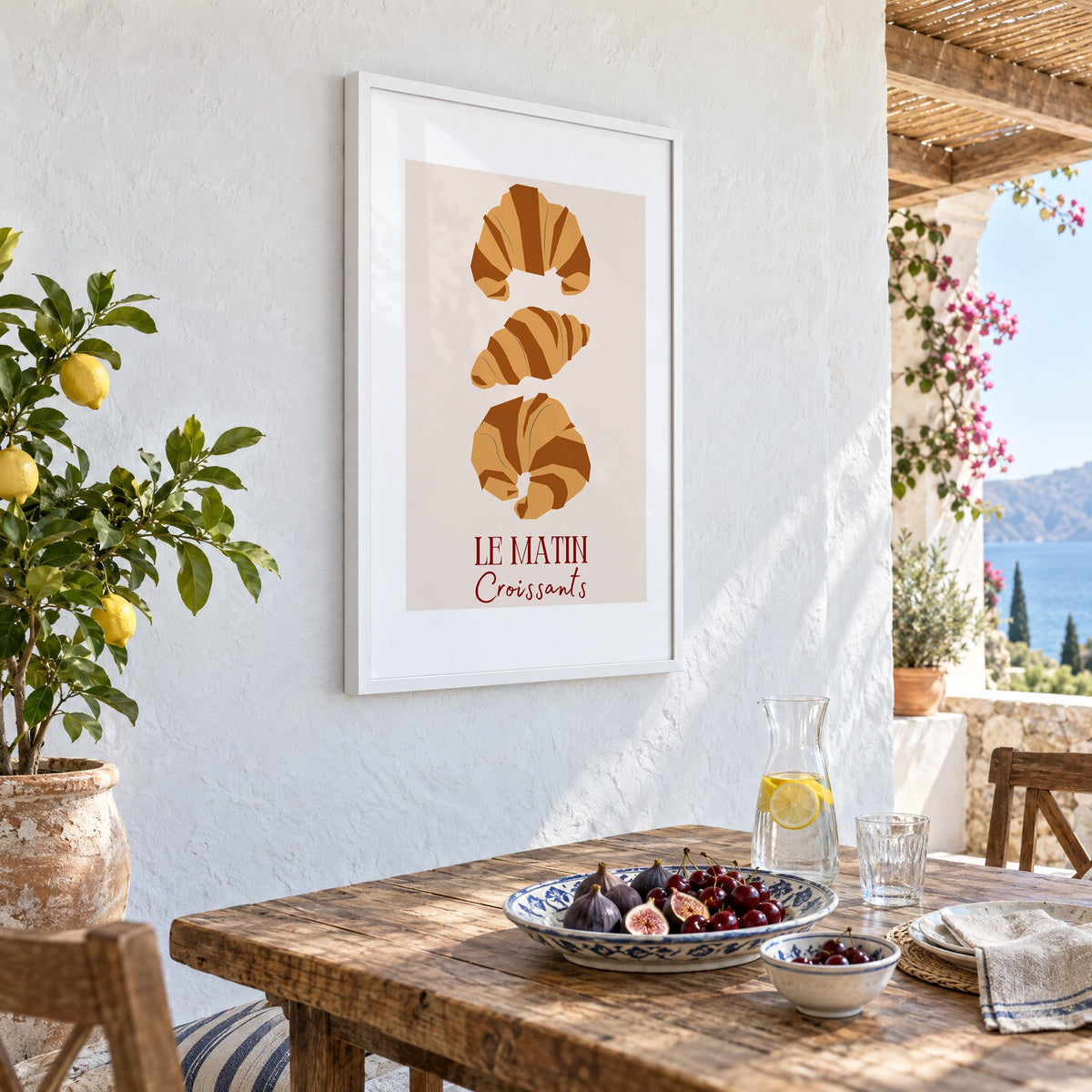 LE MATIN Croissants caramel trio print in a white frame on a whitewashed terrace wall above a lunch table with figs and cherries, seen at an angle