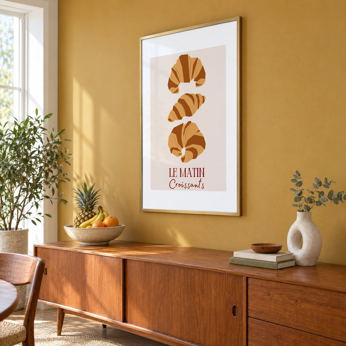 LE MATIN Croissants caramel trio print in a brass frame on a mustard wall above a teak sideboard with a fruit bowl, seen at an angle