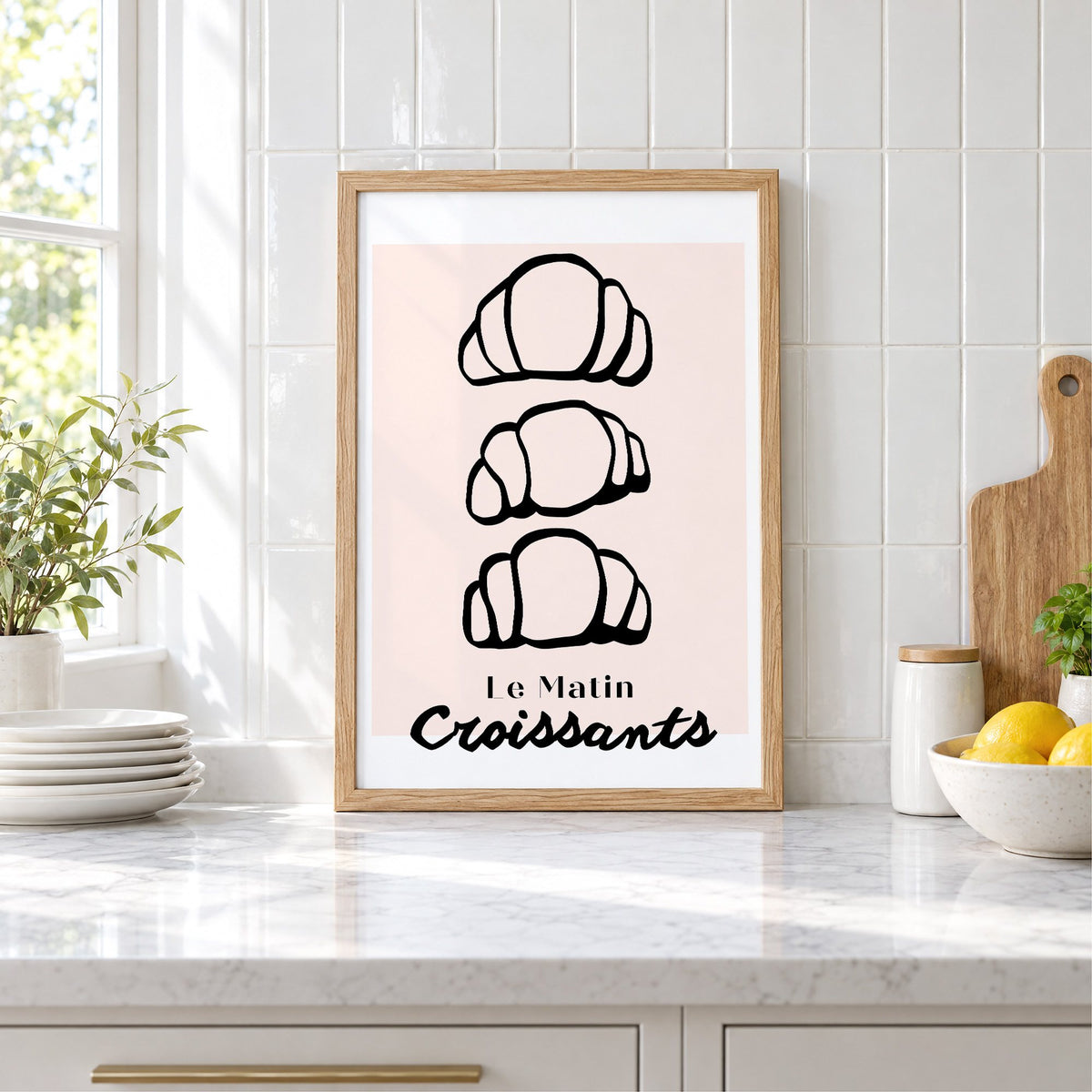 Le Matin black outline croissants print in an oak frame leaning on a marble counter by white tiles and lemons