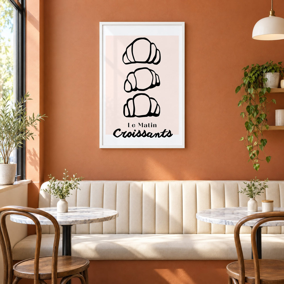 Le Matin black outline croissants print in a white frame above a cream banquette on a terracotta cafe wall