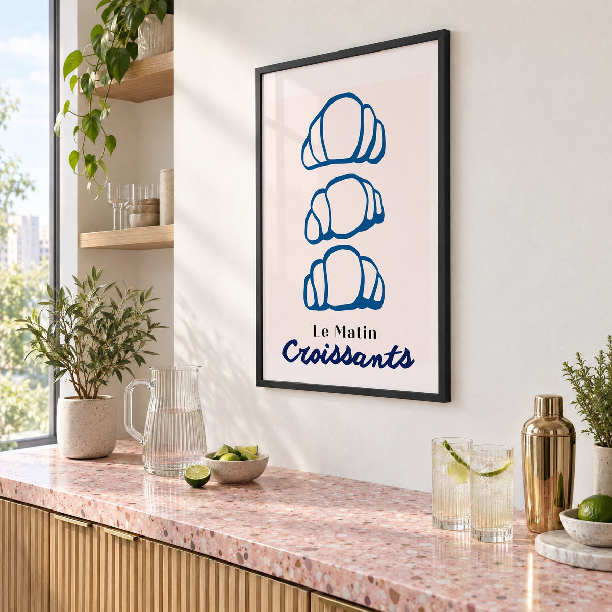 Le Matin blue croissants print in a black frame above a pink terrazzo cocktail counter with limes, seen at an angle