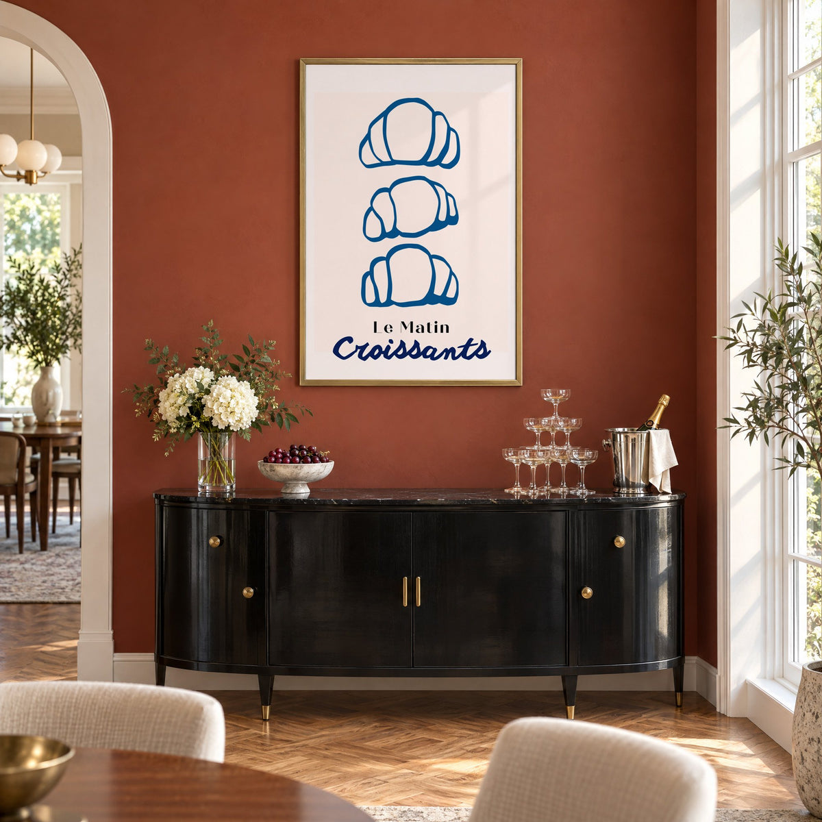 Le Matin blue croissants print in a brass frame on a terracotta wall above a black buffet with a champagne tower