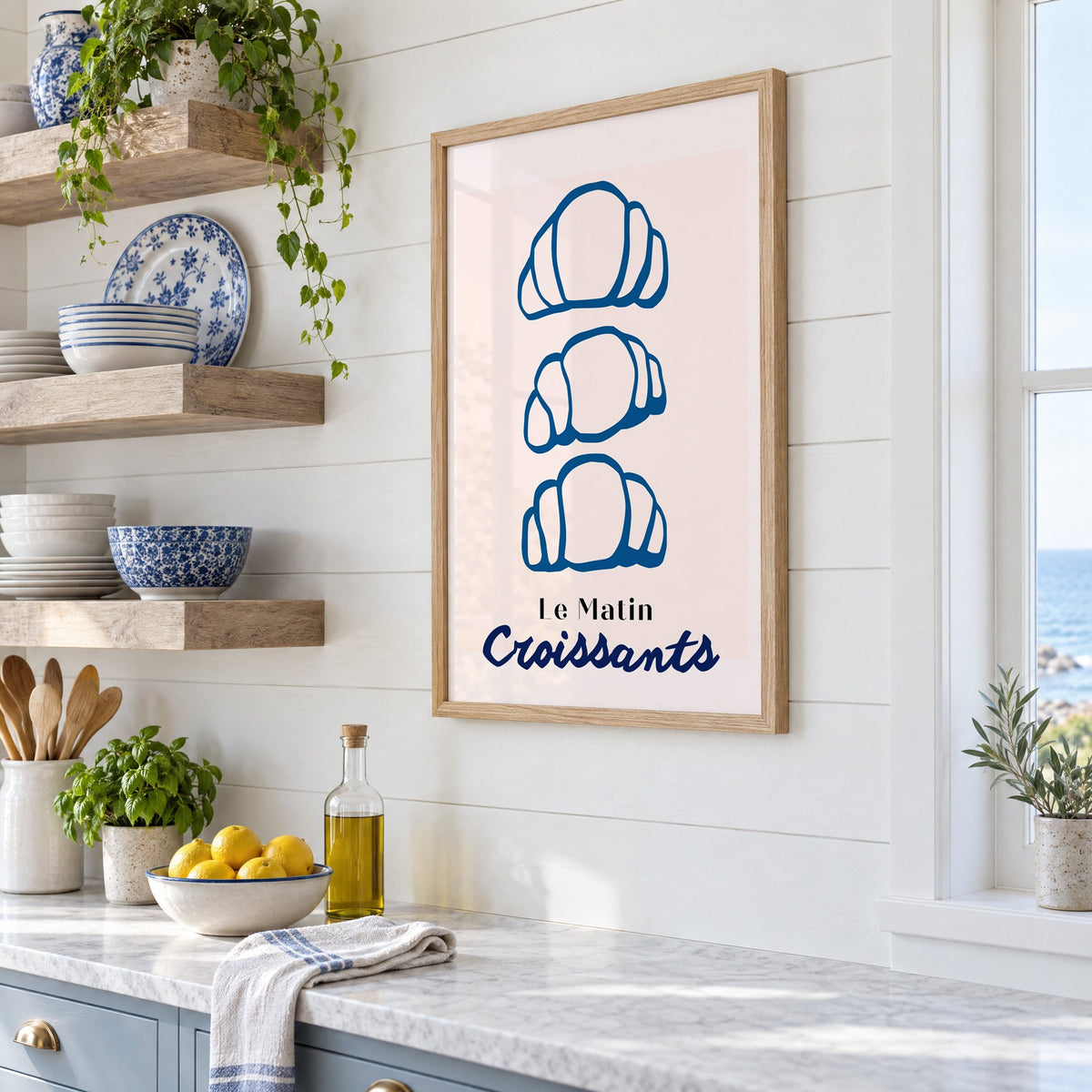 Le Matin blue croissants print in an oak frame in a white coastal kitchen beside blue and white plates