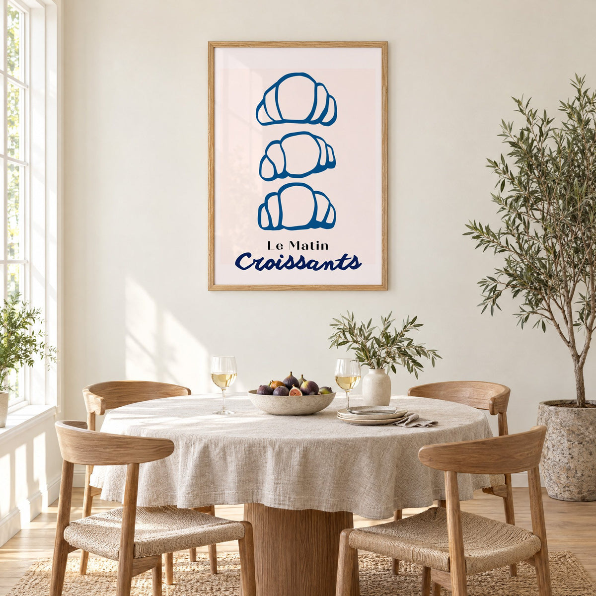 Le Matin blue croissants print in an oak frame above a round linen-covered dining table with figs and wine