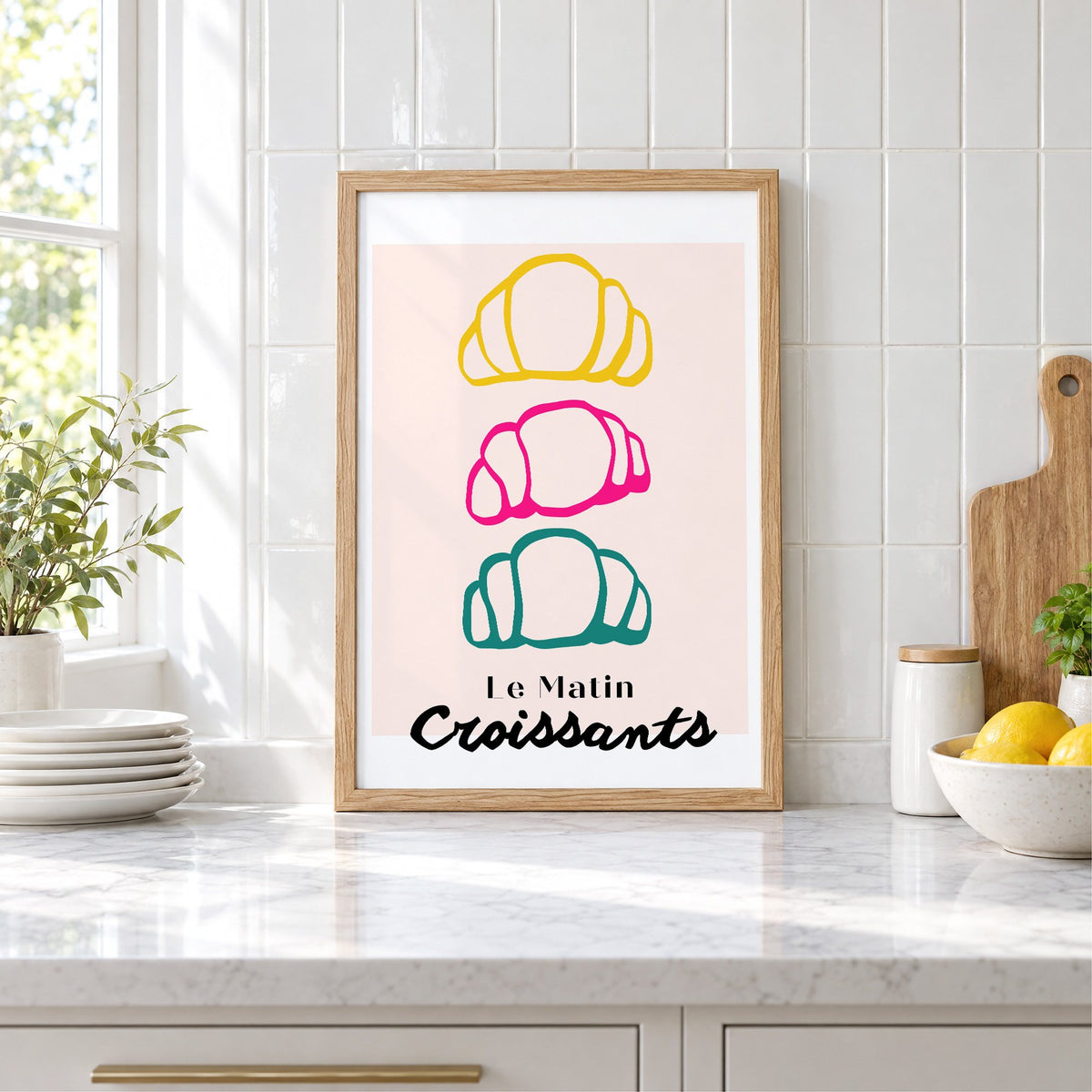 Le Matin colorful croissants print in an oak frame leaning on a marble counter by white tiles and lemons