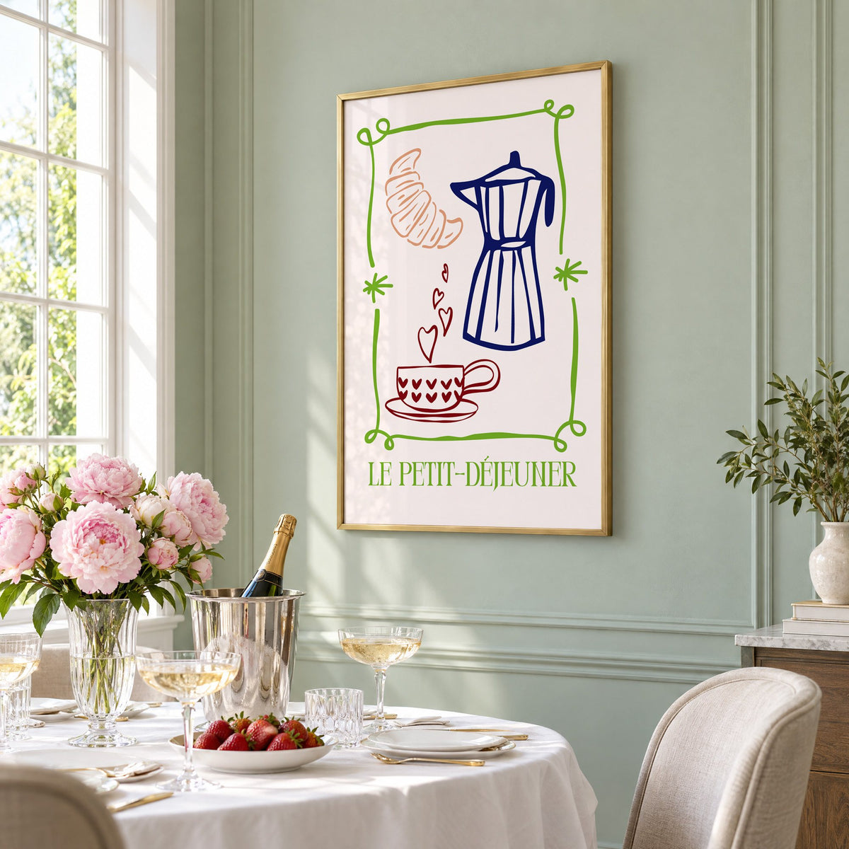 LE PETIT-DÉJEUNER moka pot and croissant print in a brass frame on a sage wall above a brunch table with champagne and peonies, seen at an angle