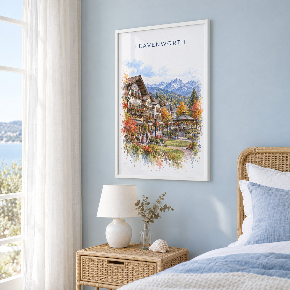 Leavenworth watercolor print in a white frame on a pale blue wall beside a rattan bed in a coastal bedroom