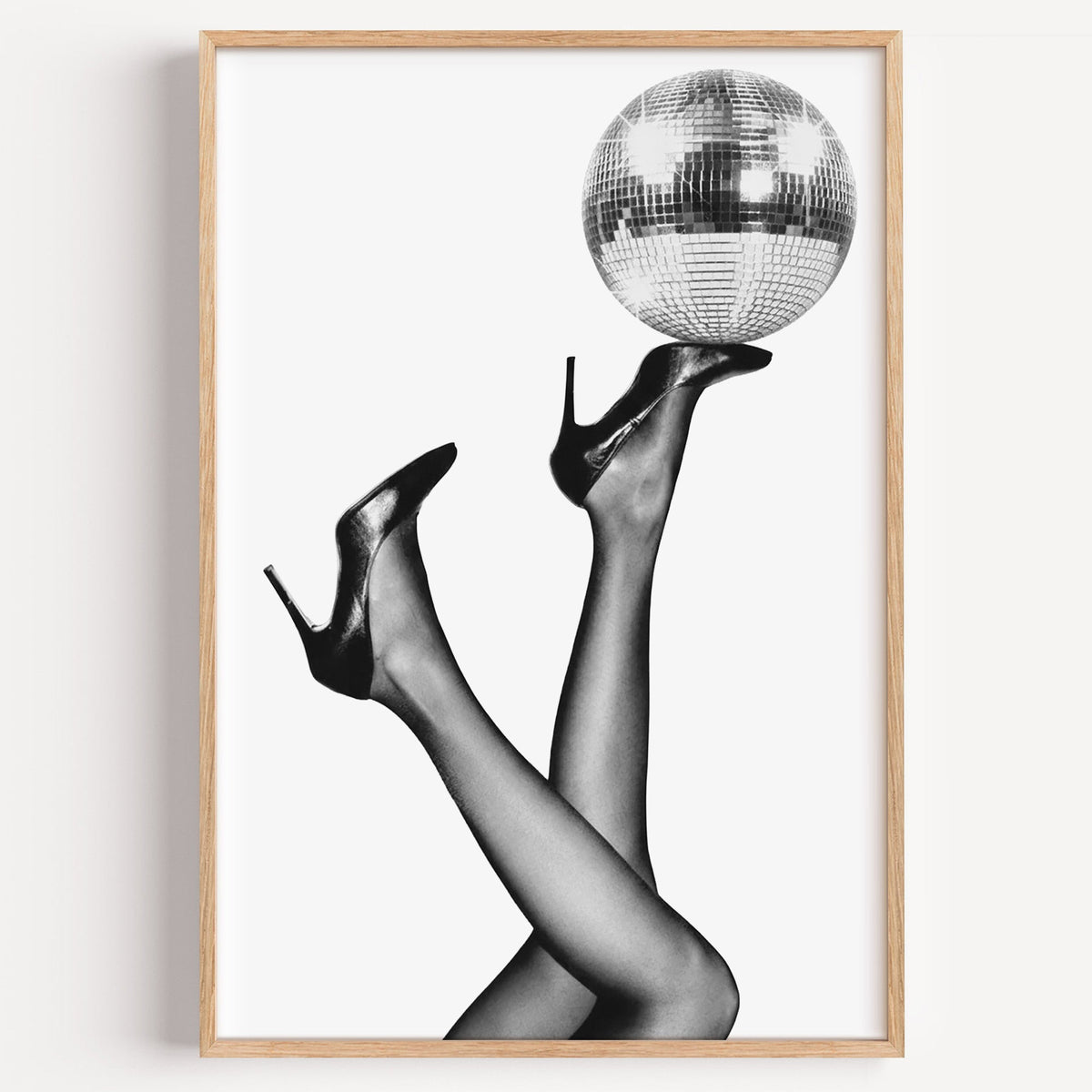Legs in heels balancing a disco ball print in a light oak frame on a plain white wall