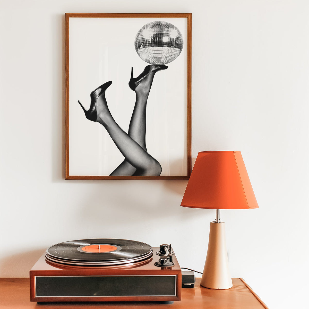 Legs in heels balancing a disco ball print in a walnut frame above a record player and orange lamp