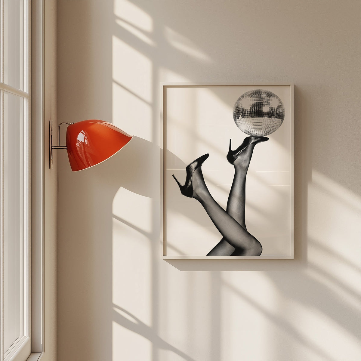 Legs in heels balancing a disco ball print in a white frame on a sunlit wall beside a red sconce