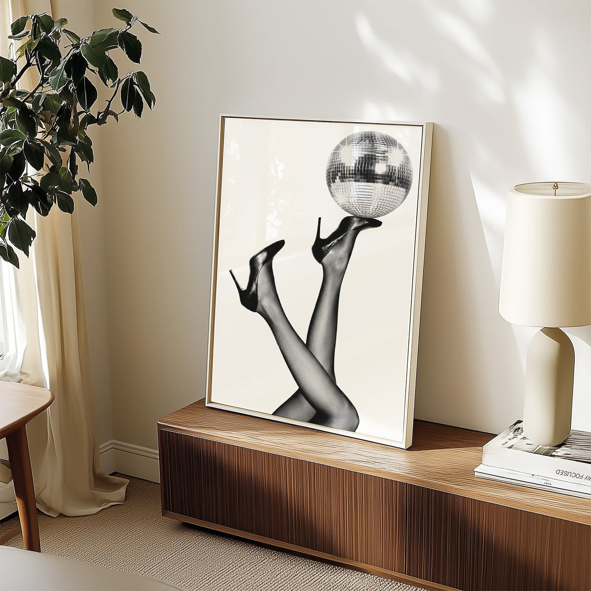 Legs in heels balancing a disco ball print in a white frame on a walnut sideboard beside a ceramic lamp