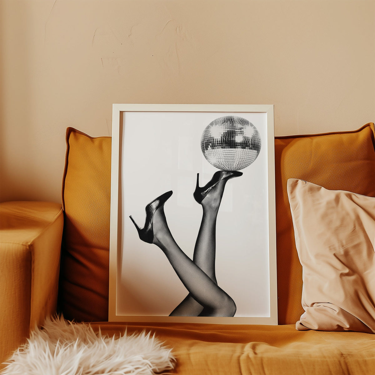 Legs in heels balancing a disco ball print in a white frame leaning on a mustard velvet sofa