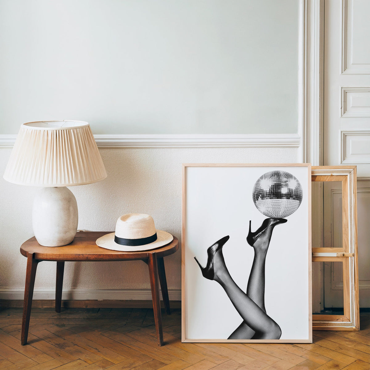 Legs in heels balancing a disco ball print in a pine frame on a wood floor beside a lamp and straw hat