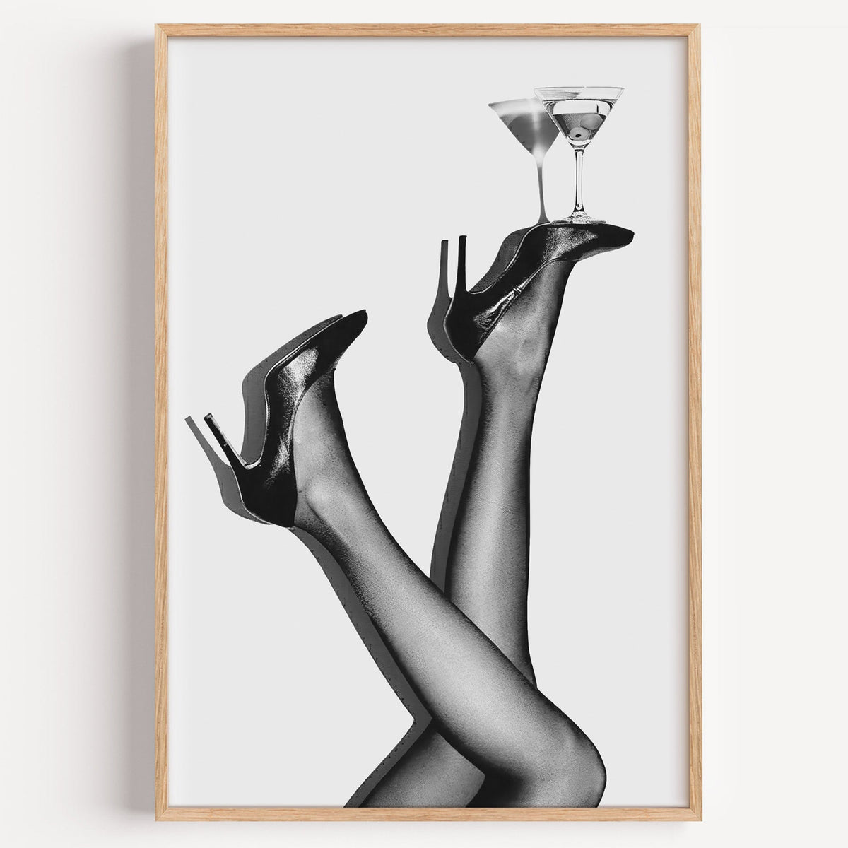 Legs in heels balancing two martini glasses print in a light oak frame on a plain white wall
